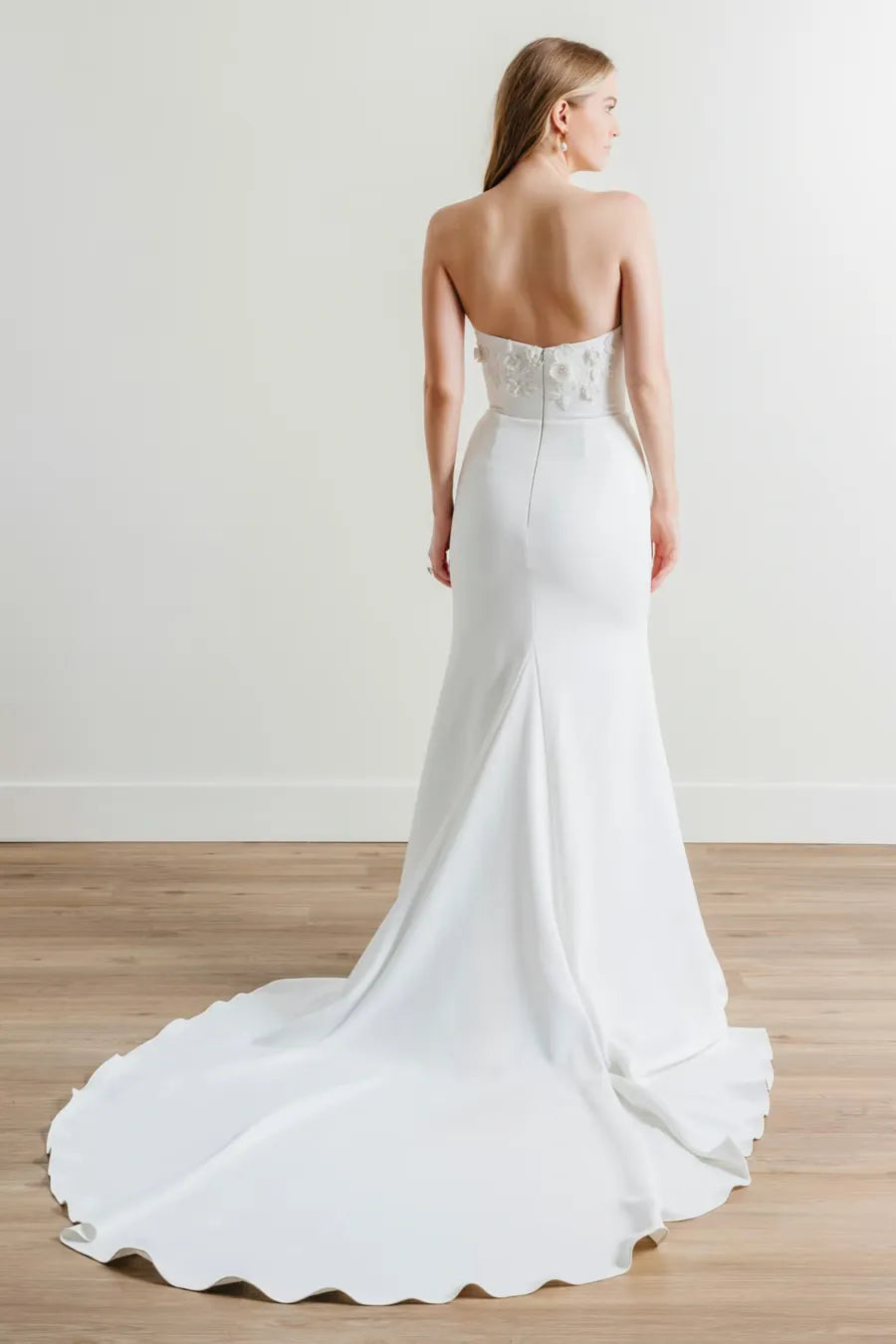 A woman wearing a strapless white wedding gown is shown from the back in a bright studio with a light wall and wood floor. The dress has a fitted silhouette, a scalloped lace-embellished back bodice, and a long flowing train.