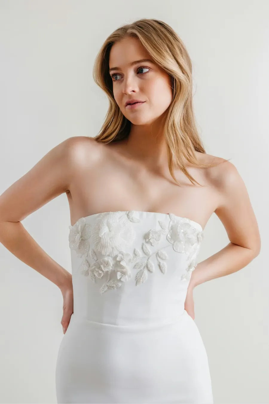 A woman modeling a strapless white dress with a structured, slightly padded bodice and 3D floral applique across the chest, set against a plain light gray studio background.