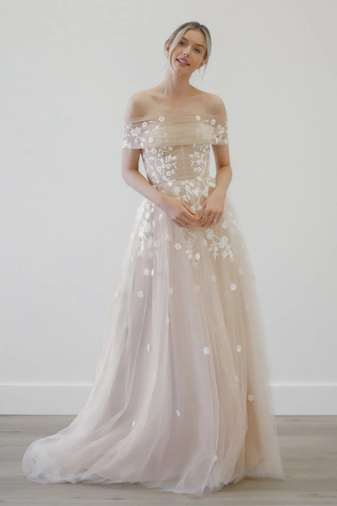 Woman wearing a beige off-shoulder gown with floral embroidery against a white background