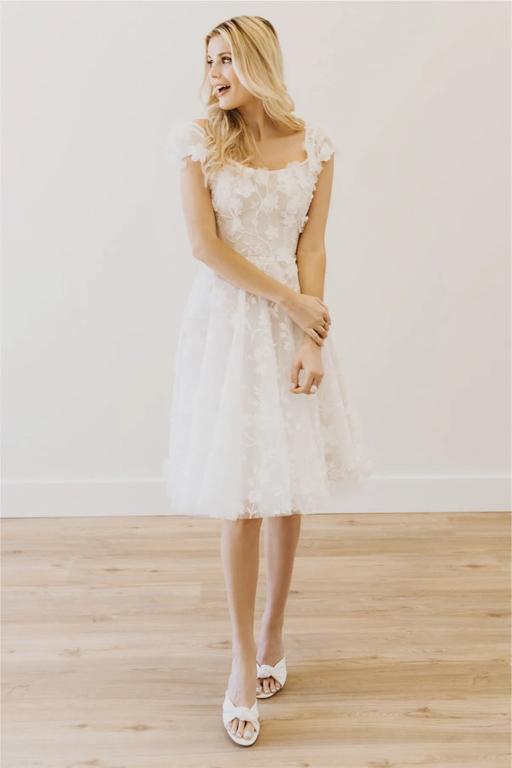 Woman wearing a white lace dress standing on a wooden floor with a plain white wall background