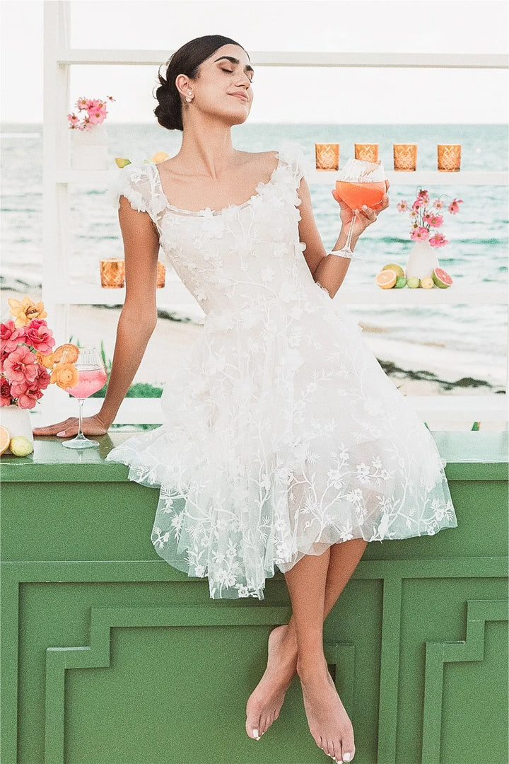 Woman in a white lace dress holding a drink at a decorated outdoor event.