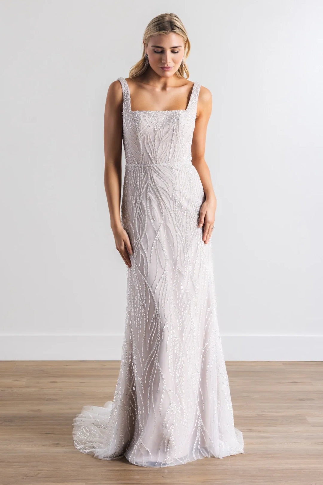 A woman modeling a sleeveless, floor-length white gown with intricate beadwork and a square neckline, standing on a light wood floor against a white wall.