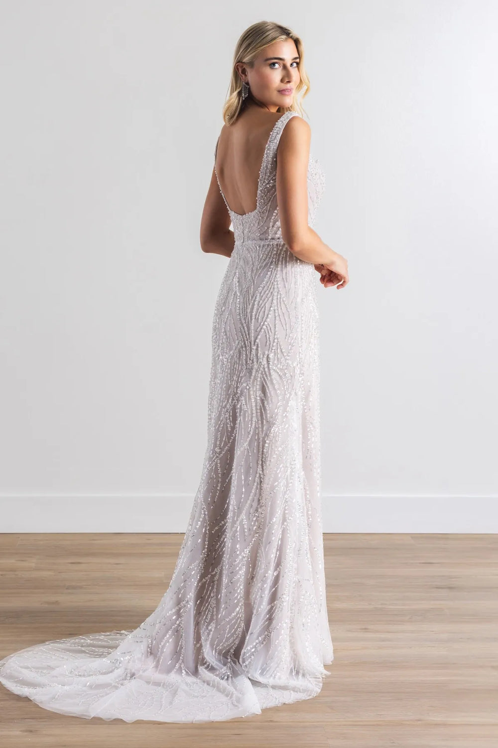 A woman modeling a sleeveless, open-back floor-length gown with intricate bead and lace detailing, standing on a light wood floor against a white wall.