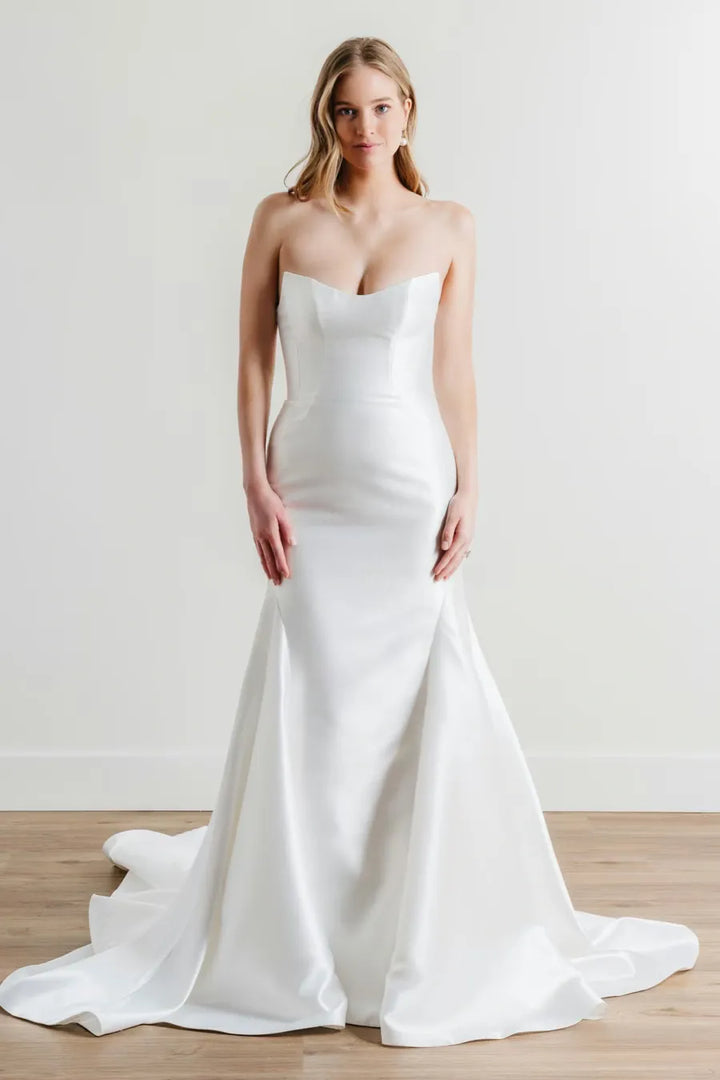 A woman modeling a strapless white satin wedding gown with a sweetheart neckline, fitted bodice, and flared mermaid-style skirt with a short train, standing against a plain light wall and wood floor.