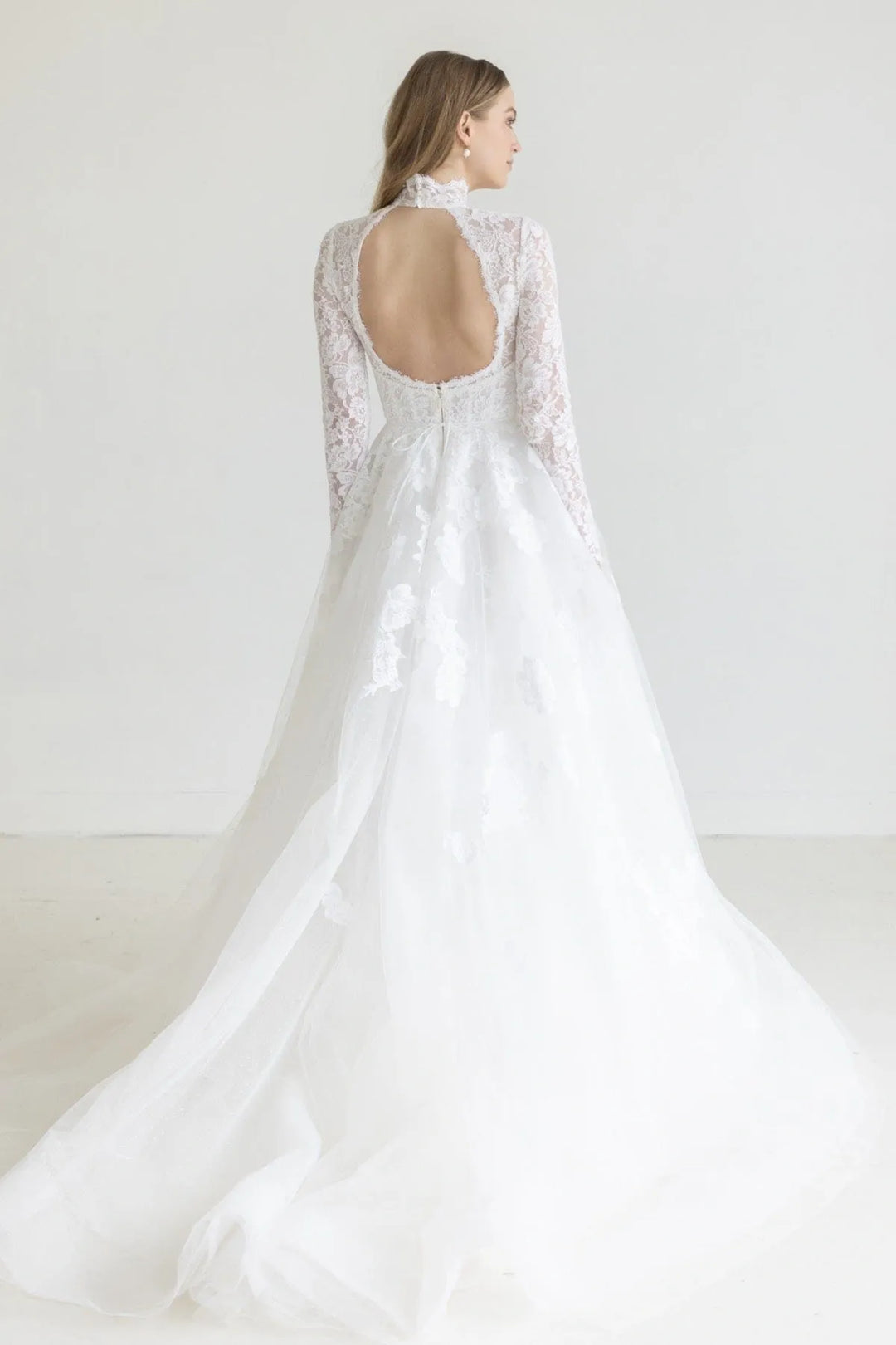 A woman wearing a white lace wedding dress with a high neck, long sleeves, and a dramatic full tulle train, shown from the back in a bright studio setting.