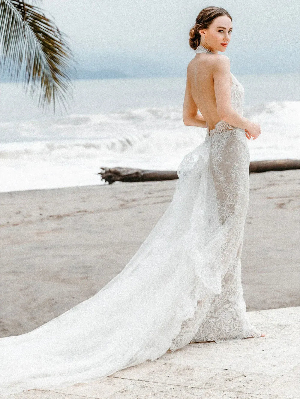 Woman in a white lace wedding dress on a beach with palm leaves.