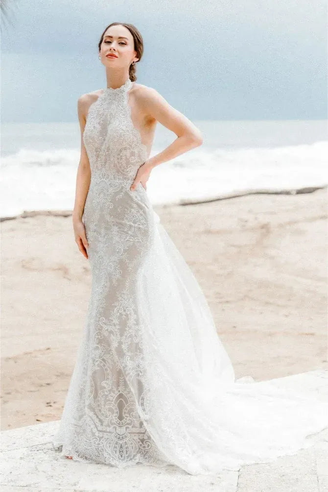 Woman in a white lace wedding dress standing on a beach.