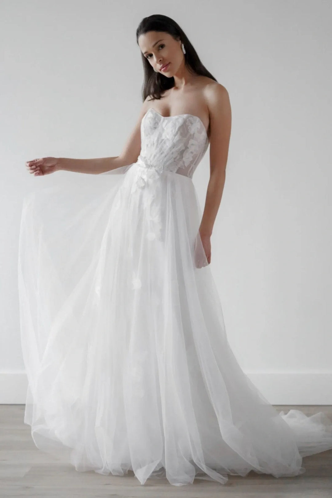 A woman modeling a strapless white wedding gown with a lace-embroidered bodice, sheer corset-style waist, and a full tulle skirt, posed in a bright studio with a light gray wall and pale wood floor.