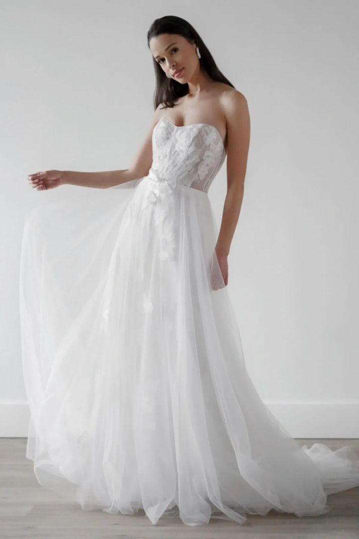 A woman modeling a strapless white wedding gown with a lace-embroidered bodice, sheer corset-style waist, and a full tulle skirt, posed in a bright studio with a light gray wall and pale wood floor.
