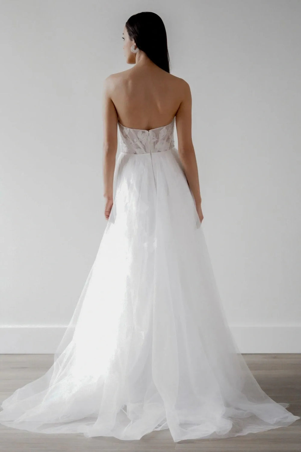 A woman shown from the back wearing a strapless white wedding gown with a sheer lace bodice and a full tulle skirt, standing against a plain light gray wall on a wood floor.