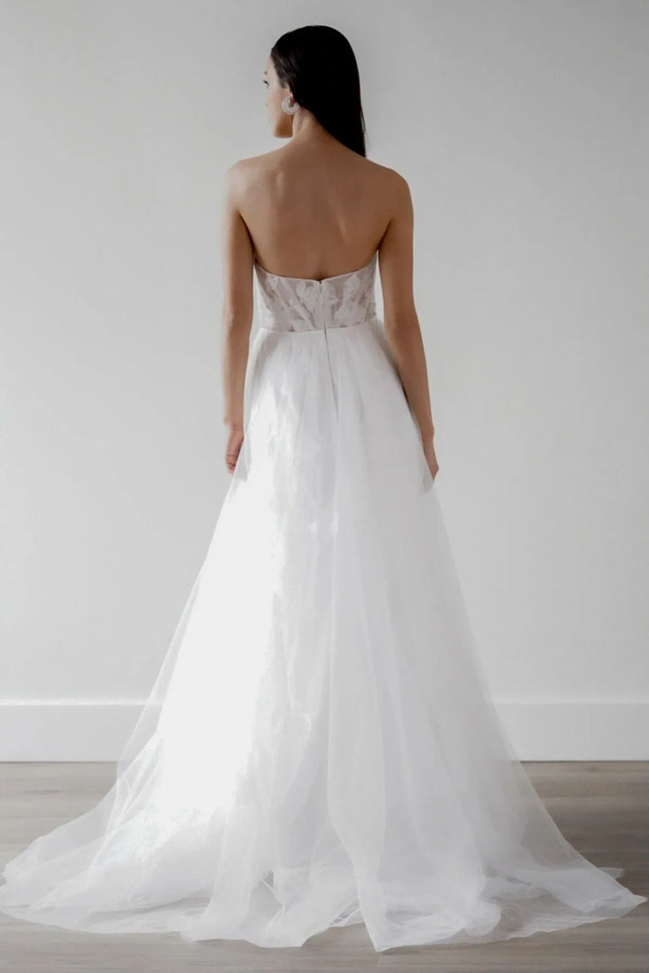 A woman shown from the back wearing a strapless white wedding gown with a sheer lace bodice and a full tulle skirt, standing against a plain light gray wall on a wood floor.