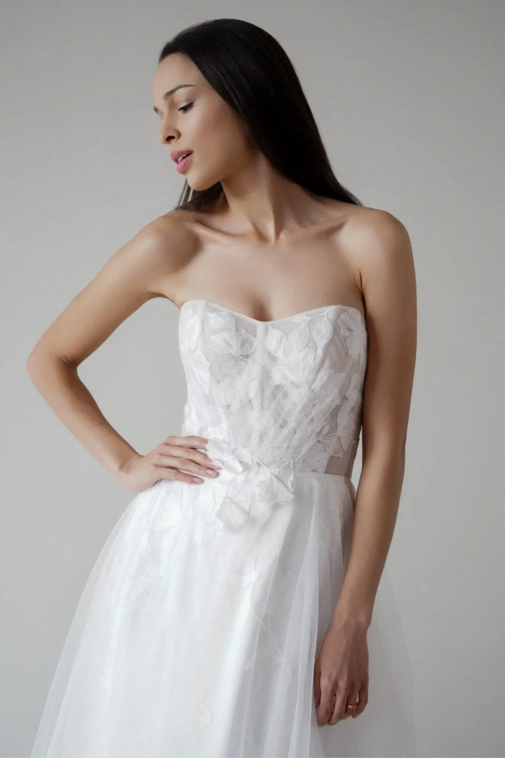 A woman modeling a strapless white wedding dress with a lace-embroidered bodice, sheer side panels, and a full tulle skirt against a plain light gray studio background.
