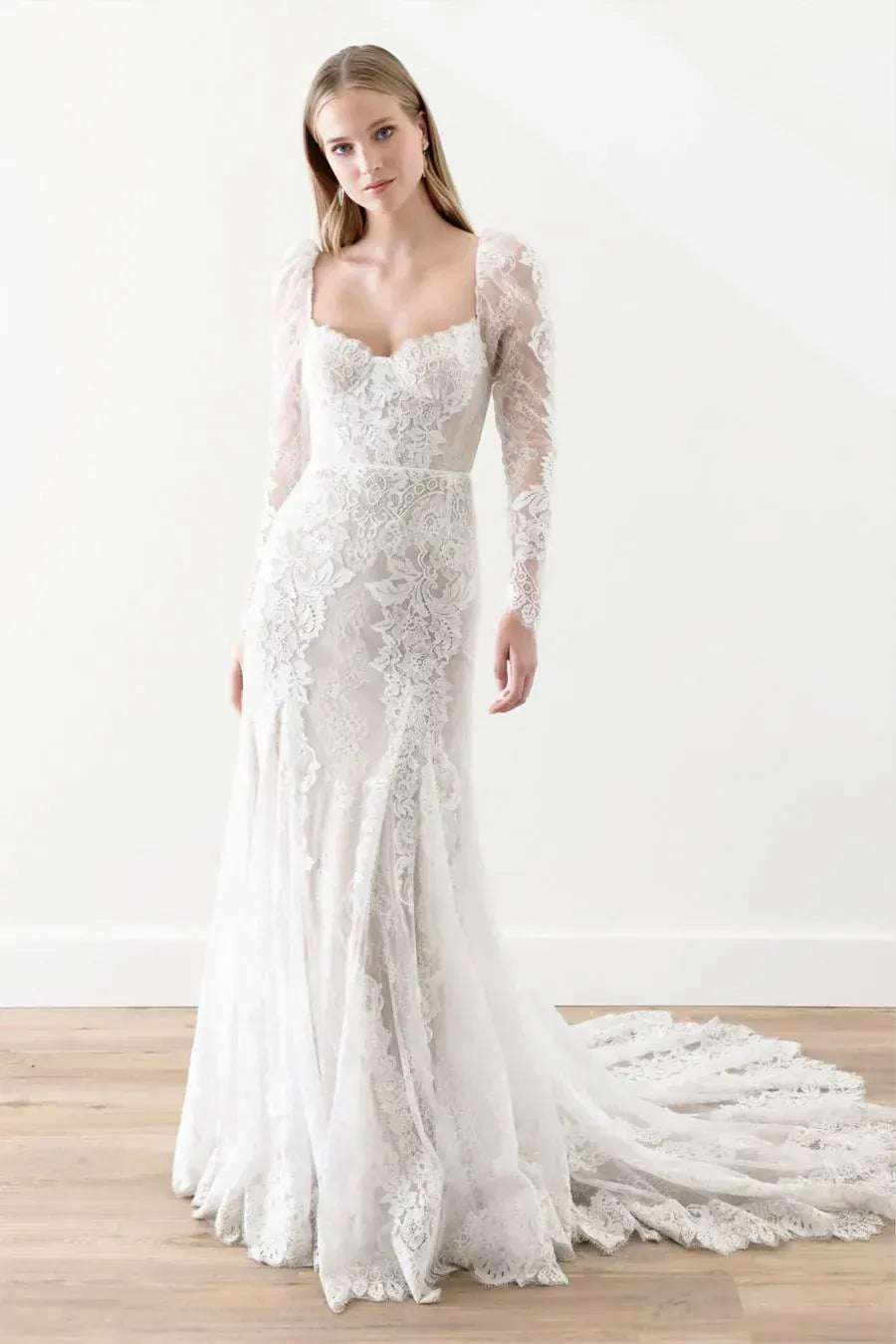 A woman modeling a long-sleeve white lace wedding gown with a fitted bodice, sheer sleeves, and a full skirt with a train, standing in a bright indoor studio with a light wood floor.