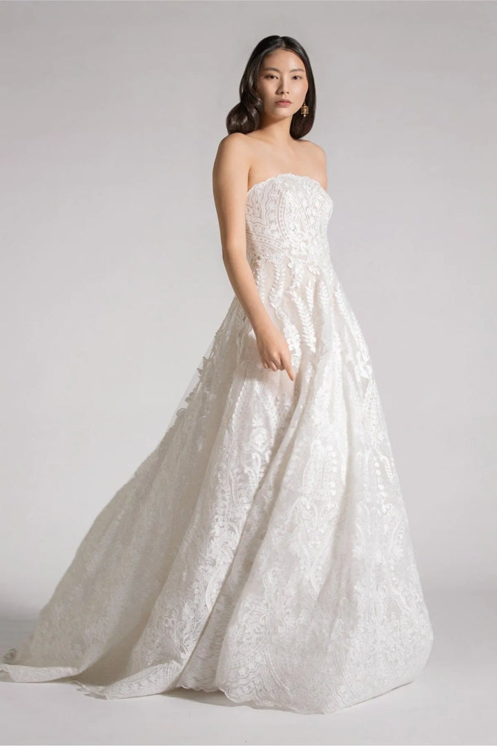 A woman modeling a strapless white lace wedding gown with a fitted bodice and full, flowing skirt against a plain light gray studio background.