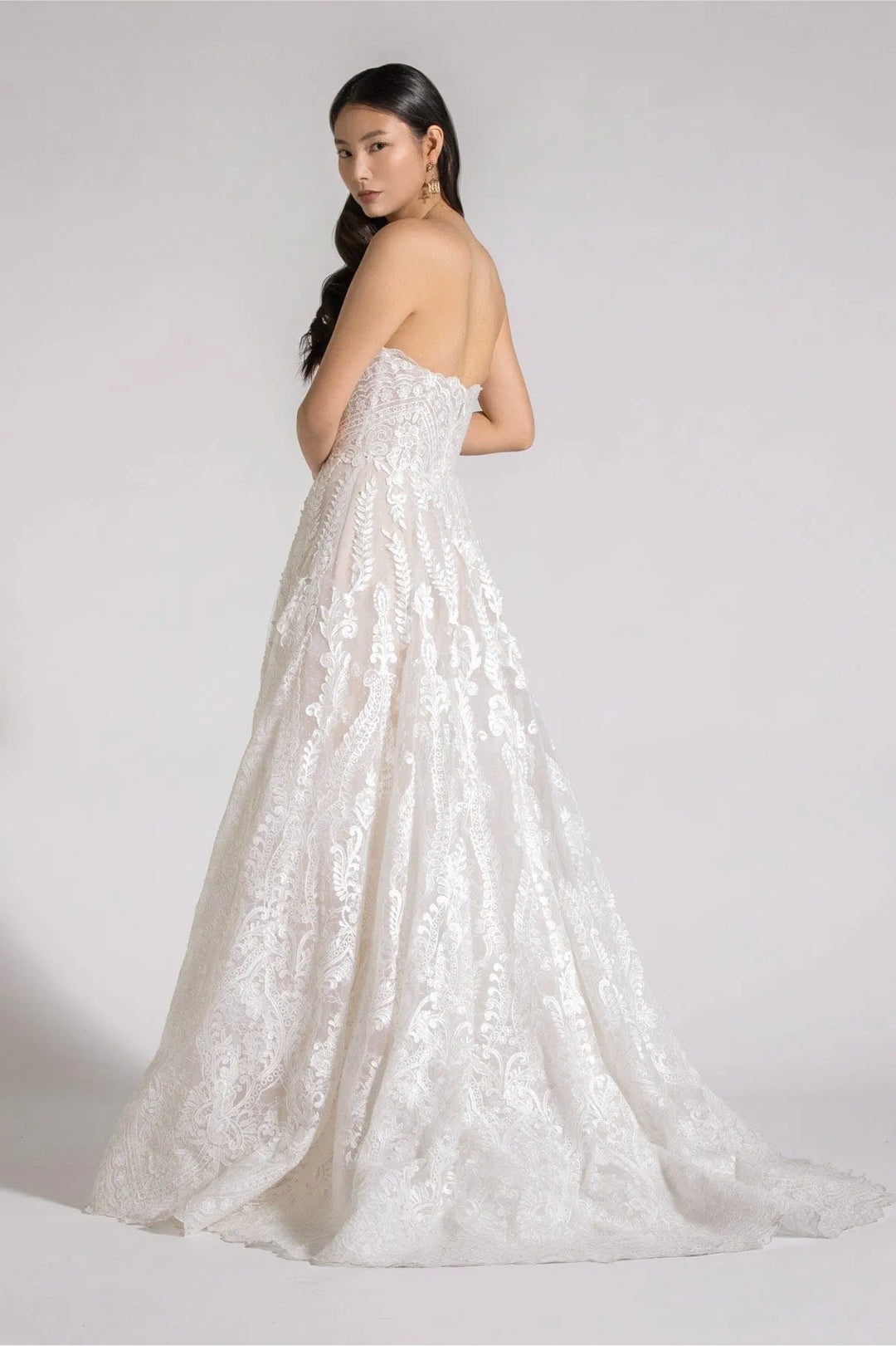 A woman modeling a strapless white lace wedding gown from the back, showing a full A-line skirt with a slight train, against a plain light gray studio background.