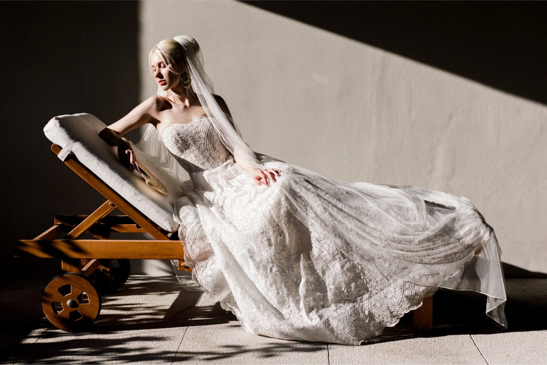 A woman in a white lace wedding gown and veil reclines on a wooden lounge chair in a sunlit interior with dramatic shadows.