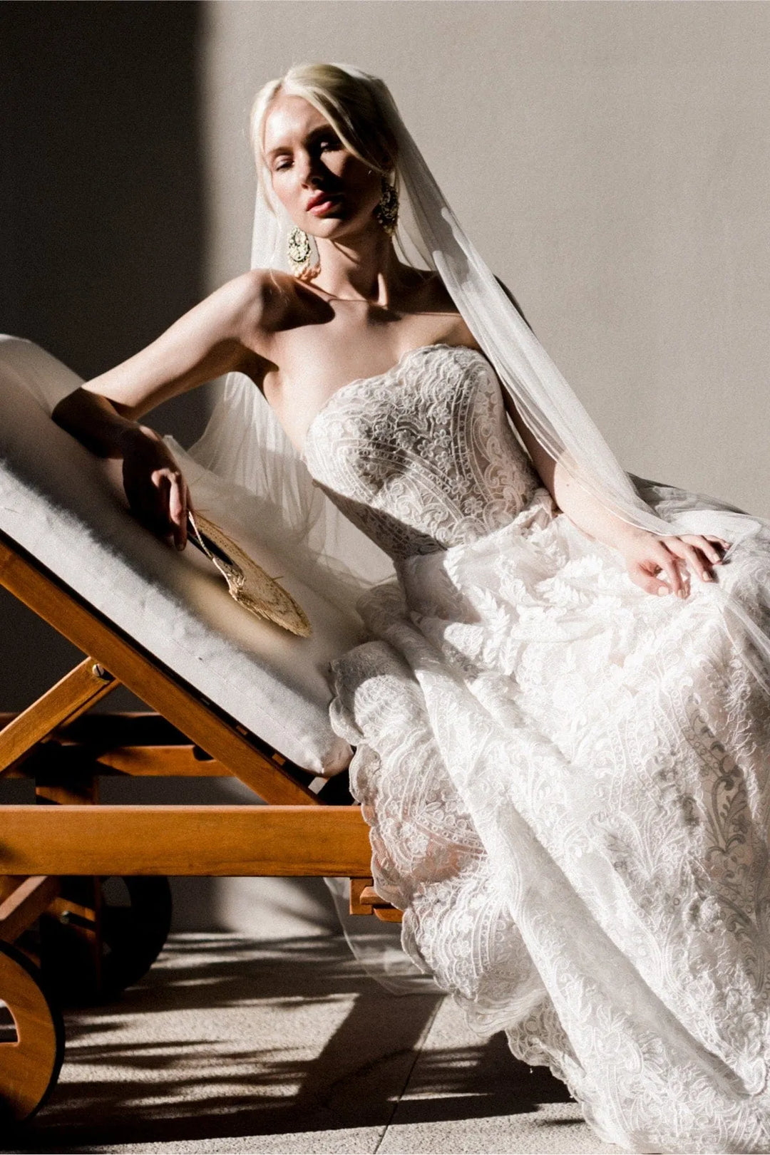 A woman wearing a strapless white lace wedding gown and long sheer veil reclines on a wooden lounge chair in dramatic side lighting against a neutral wall.
