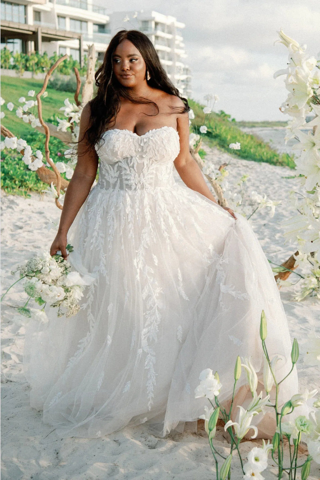 Woman in a white wedding dress standing on a beach with flowers and buildings in the background