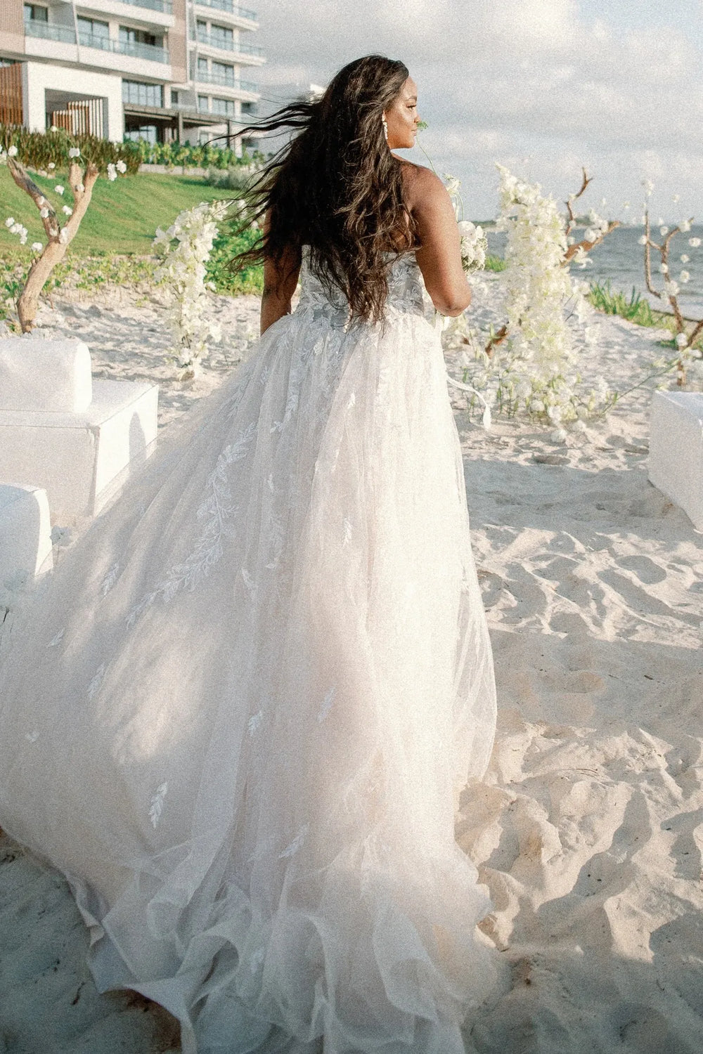 Woman in a white wedding dress on a beach with a building and greenery in the background