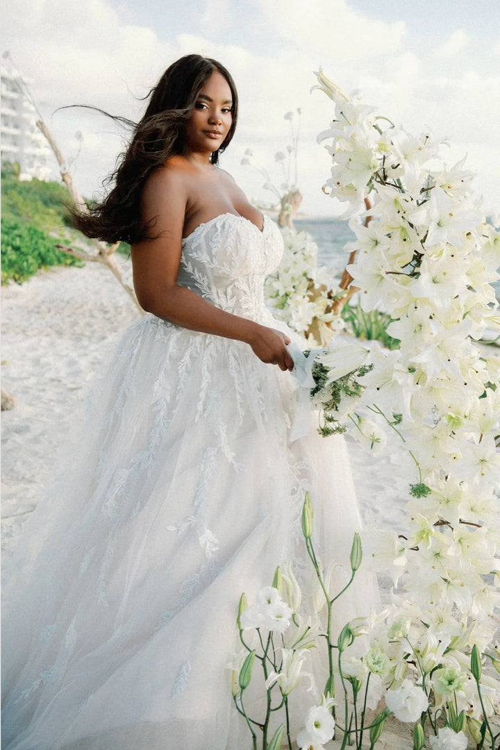 Woman in a white wedding dress standing next to a floral arch on a beach.
