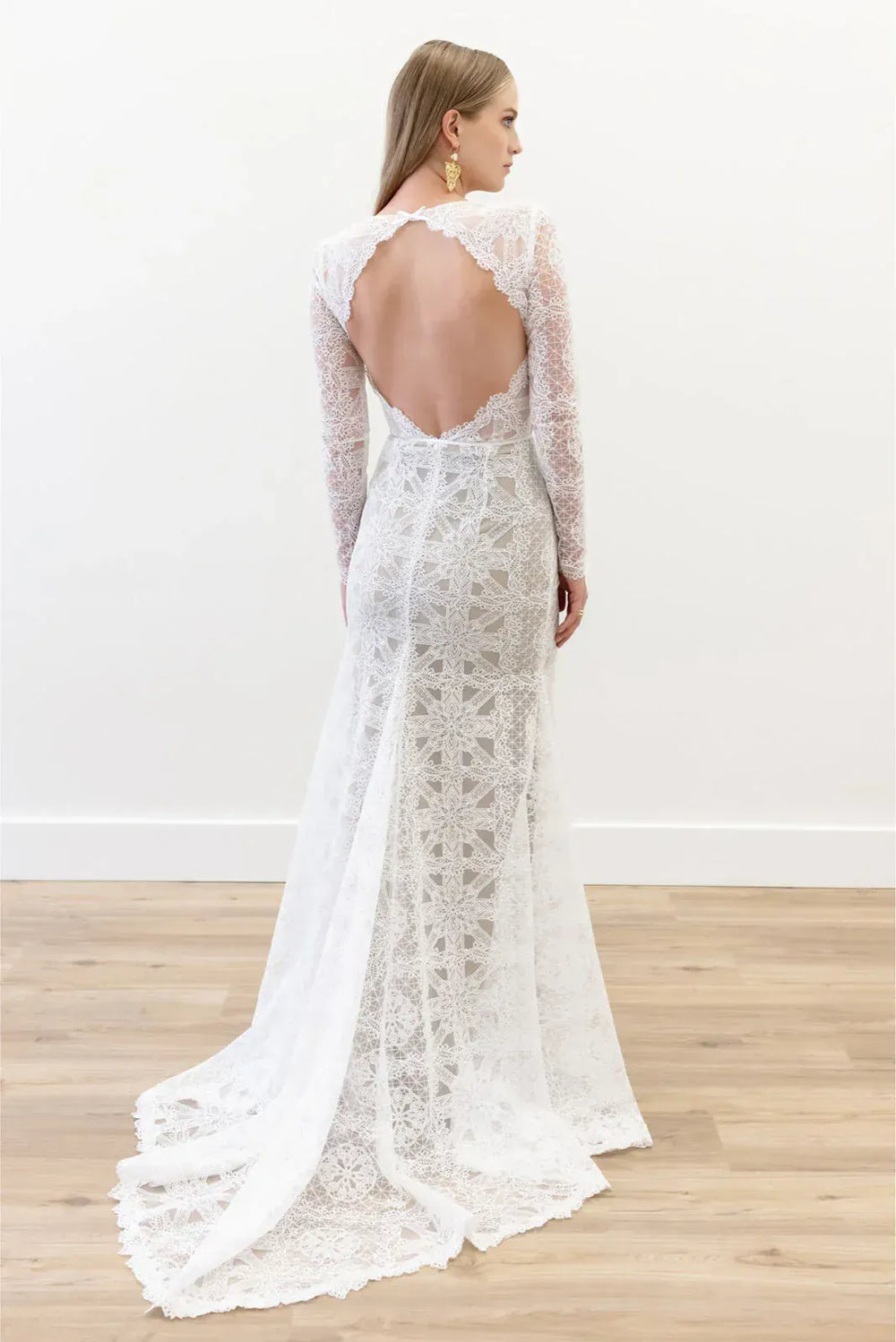 Woman wearing a white lace wedding dress with long sleeves and an open back, standing on a wooden floor.