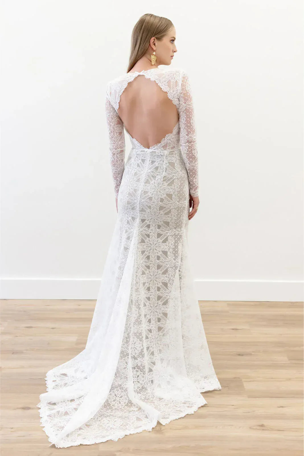 Woman wearing a white lace wedding dress with long sleeves and an open back, standing on a wooden floor.