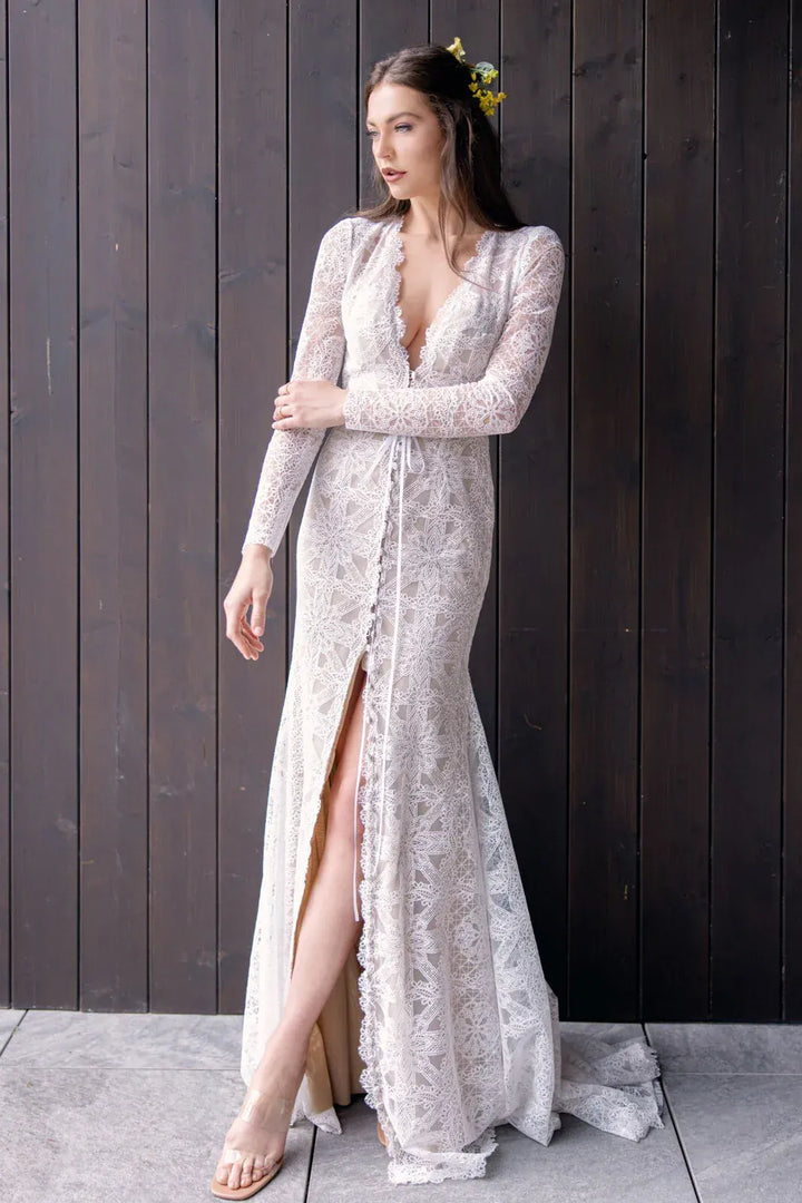 Woman wearing a lace dress with long sleeves and a high slit, standing against a wooden wall.