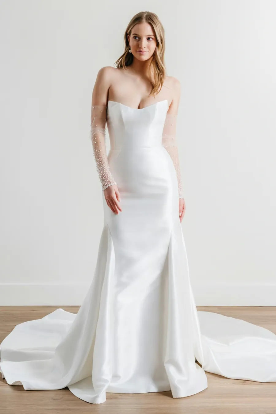 A woman modeling a strapless white satin wedding gown with sheer lace long sleeves, standing in a bright studio with a light wood floor and white wall.