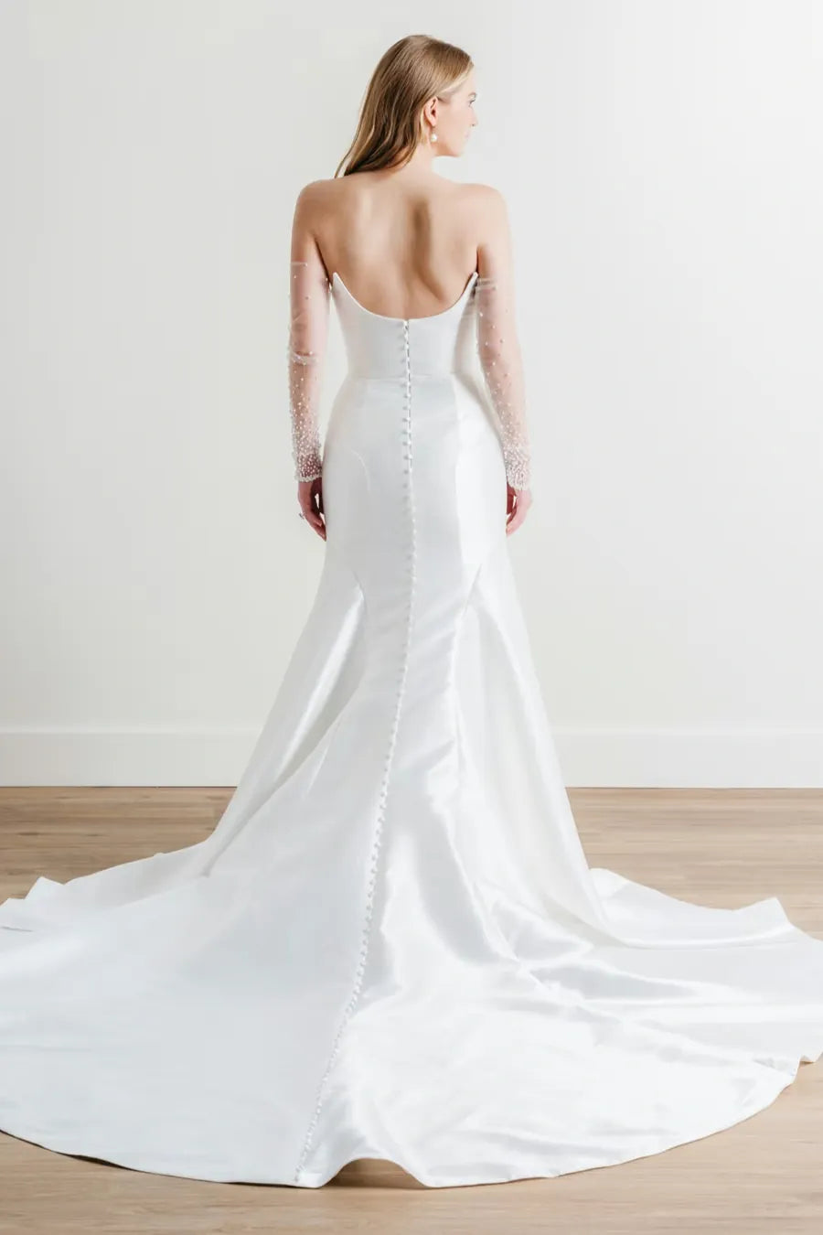 A woman shown from the back wearing a white bridal gown with a long satin train, open back, lace sleeves, and a central lace-up detail, photographed in a bright indoor studio setting.