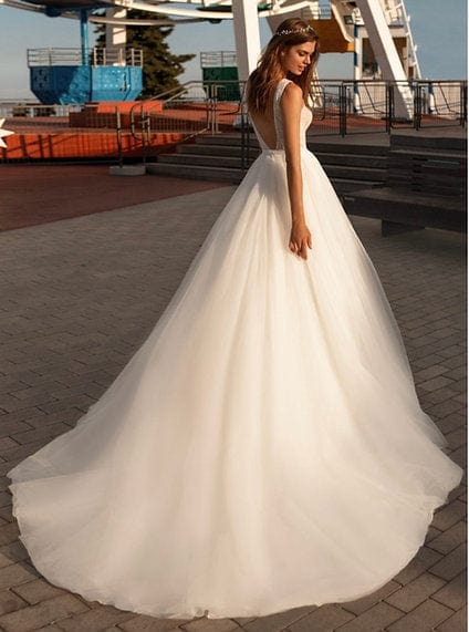A woman wearing a voluminous white wedding gown with a fitted bodice and long tulle train, standing outdoors on a paved walkway near a waterfront structure.