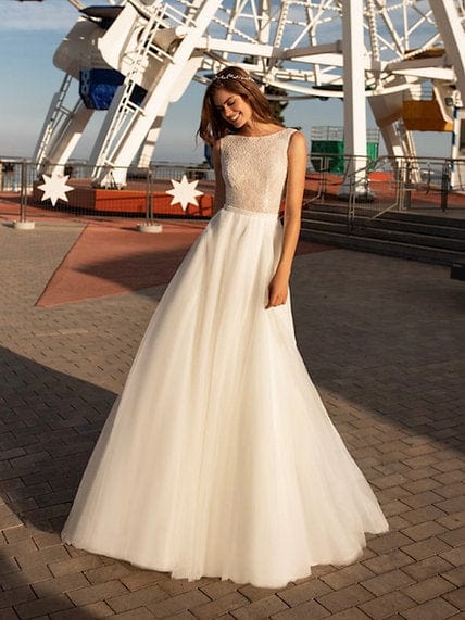 A woman wearing a sleeveless A-line wedding dress with a beaded bodice and full tulle skirt, standing outdoors on a paved walkway with a large white ferris wheel in the background.