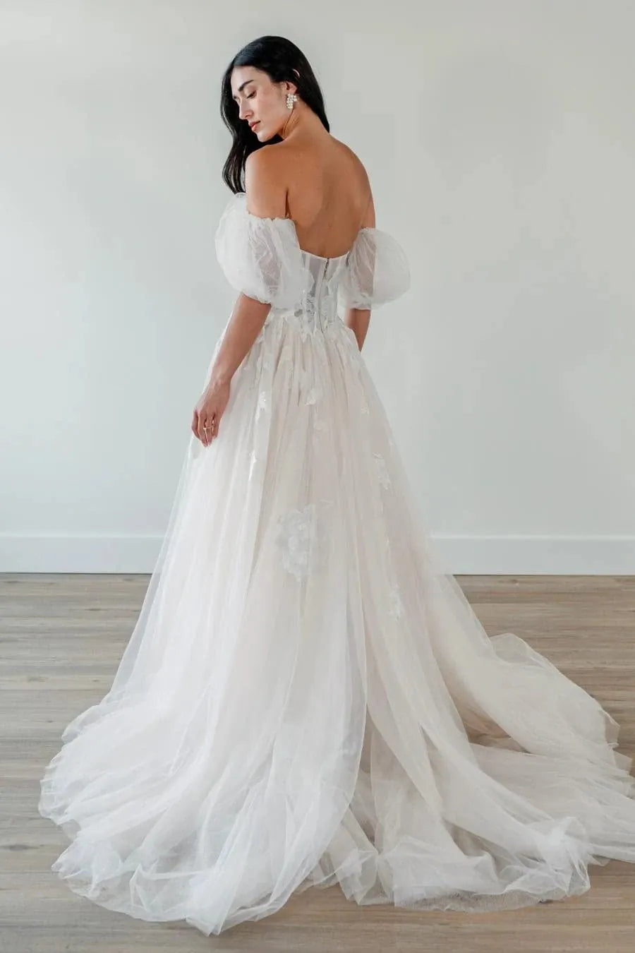 A woman wearing a voluminous off-the-shoulder tulle wedding gown with a sheer corset-style bodice and a long train, photographed in a minimal indoor room with light walls and wood flooring.