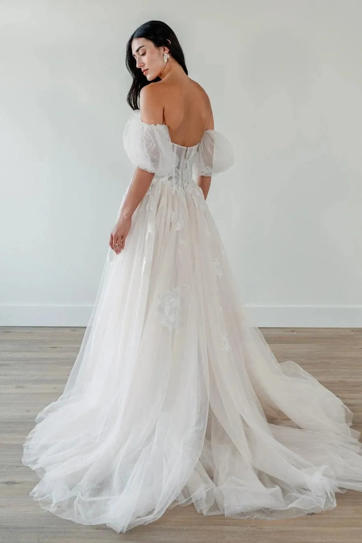 A woman wearing a voluminous off-the-shoulder tulle wedding gown with a sheer corset-style bodice and a long train, photographed in a minimal indoor room with light walls and wood flooring.