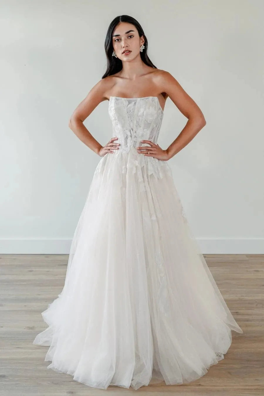 A woman modeling a strapless white wedding gown with a fitted lace corset bodice and a voluminous tulle skirt, standing in a bright indoor room with a light wall and wood floor.