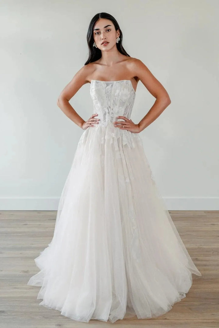 A woman modeling a strapless white wedding gown with a fitted lace corset bodice and a voluminous tulle skirt, standing in a bright indoor room with a light wall and wood floor.