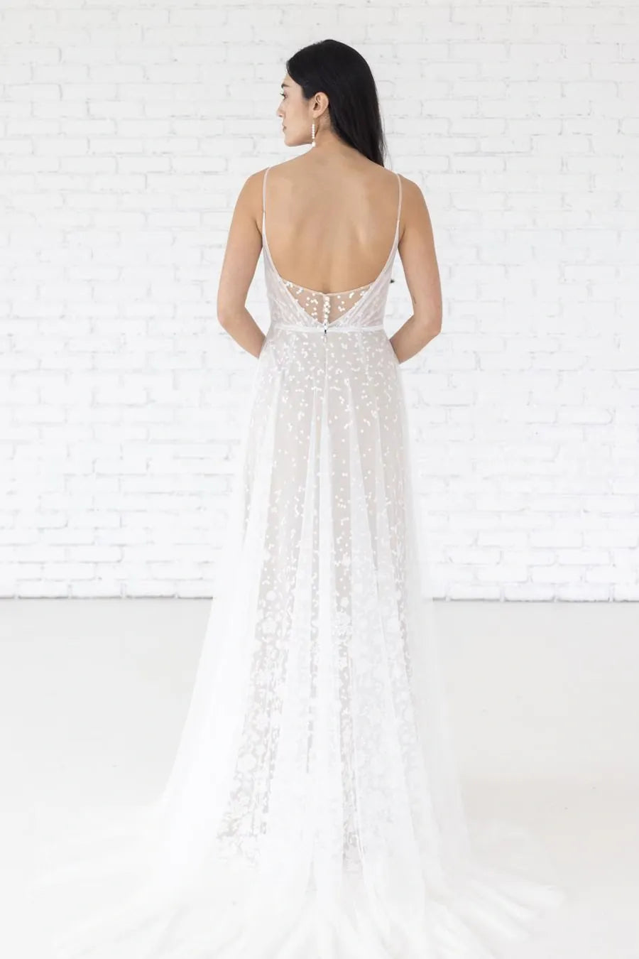 Woman wearing a white lace dress with an open back against a white brick wall.