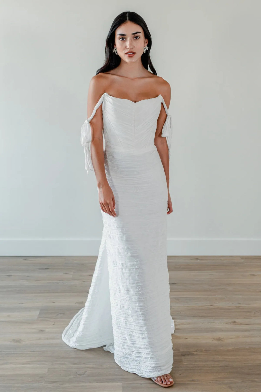 A woman modeling an off-the-shoulder white textured gown with a fitted bodice, flowing skirt, and train, standing on a light wood floor against a plain light gray wall.