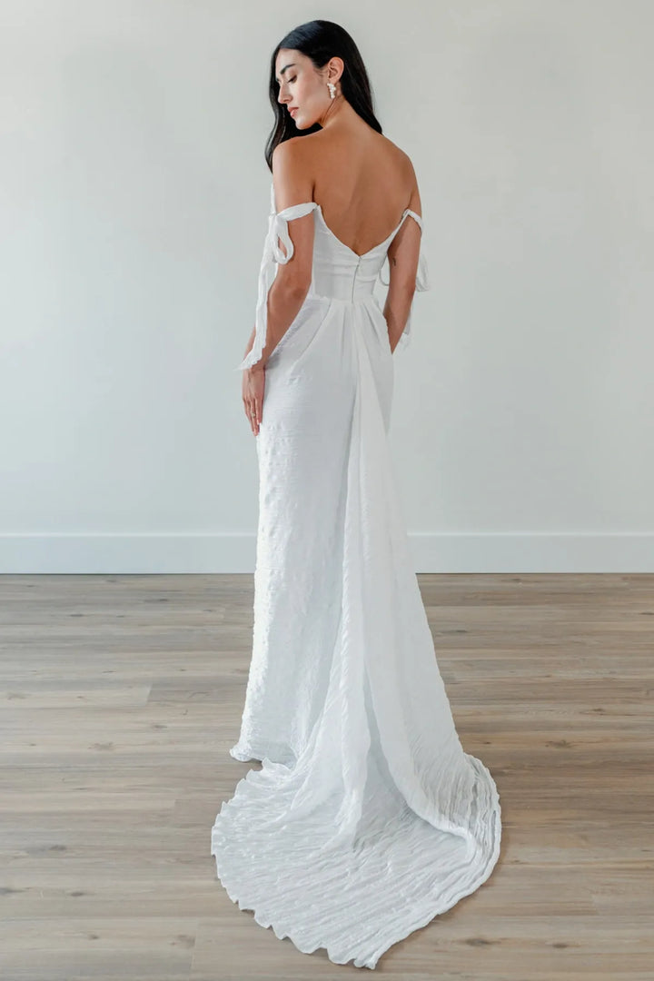 A woman modeling a white off-the-shoulder wedding gown from the back, showing a fitted bodice, open back, and long flowing train, standing on a light wood floor against a plain light wall.