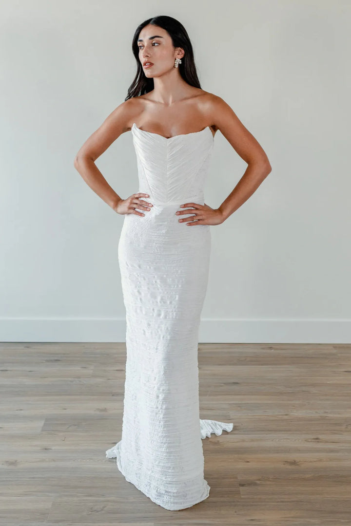 A woman modeling a strapless white mermaid-style gown with a textured, crinkled fabric, standing on a light wood floor against a neutral off-white wall.