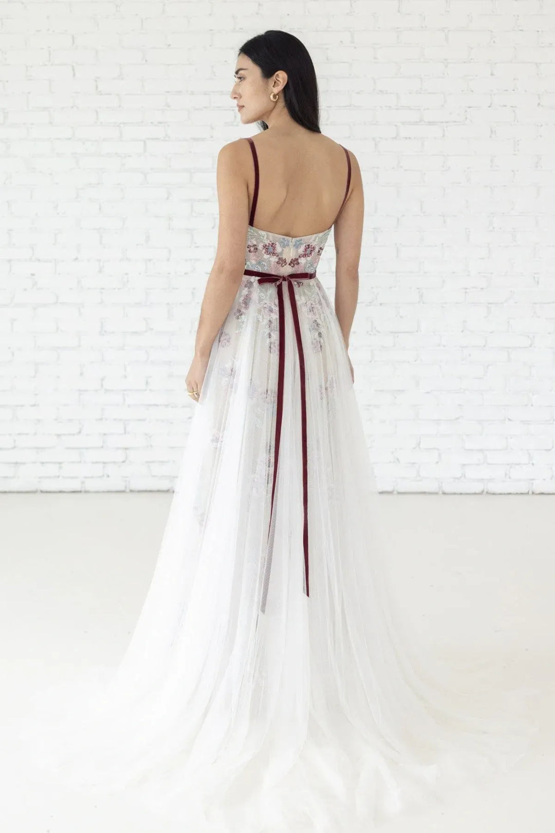 Woman wearing a white dress with floral patterns and a red belt against a white brick wall.
