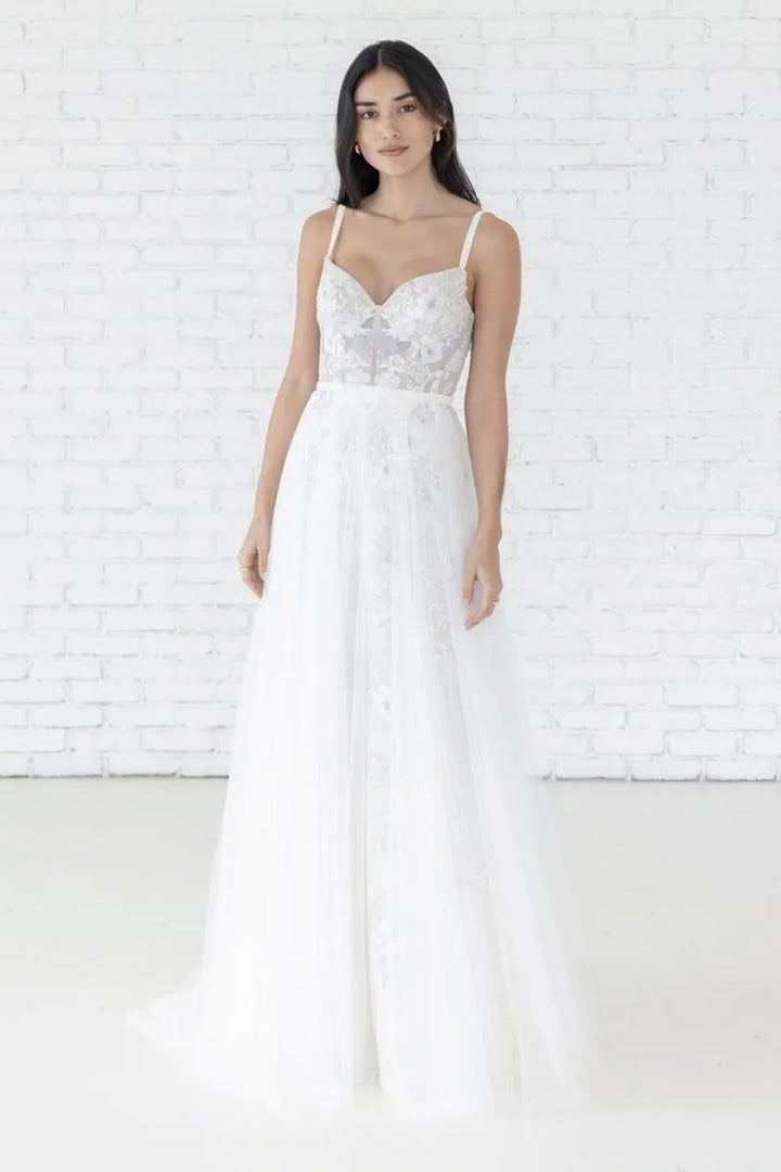 Woman wearing a white wedding dress with floral details against a white brick wall.