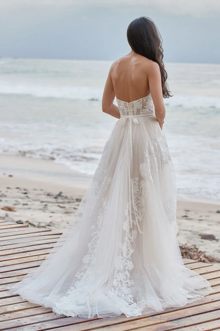 A woman stands on a wooden boardwalk by the beach wearing a strapless white lace wedding gown, viewed from behind as she faces the ocean.