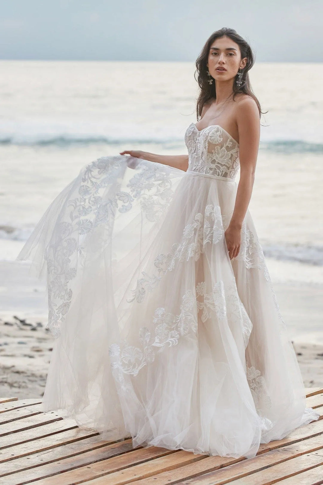 A woman wearing a strapless lace wedding gown stands on a wooden boardwalk by the beach, holding out the full train of the dress. The gown has a fitted lace bodice and a layered tulle skirt with floral lace appliqué.