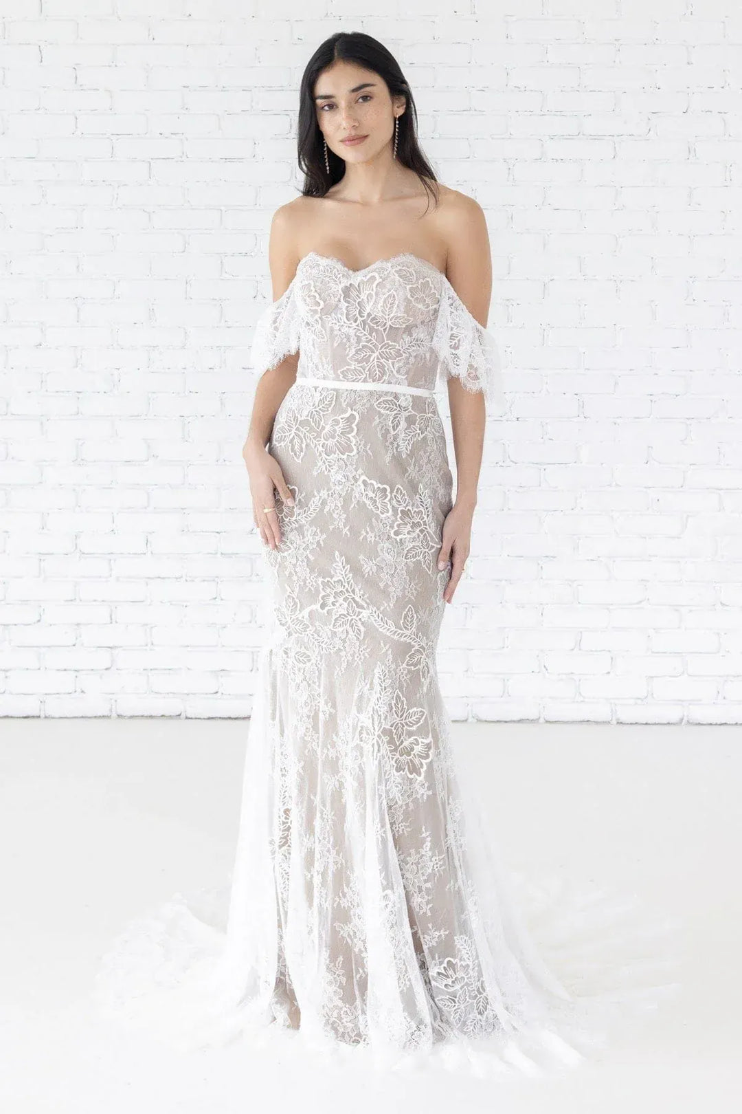 Woman wearing a white lace dress against a white brick wall.