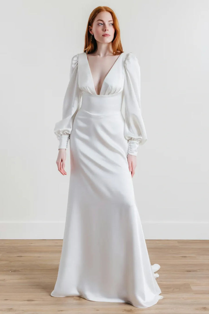 A woman modeling a floor-length white satin gown with a deep V-neckline, puffed long sleeves, and a fitted silhouette, standing against a plain light wall on a wood floor.