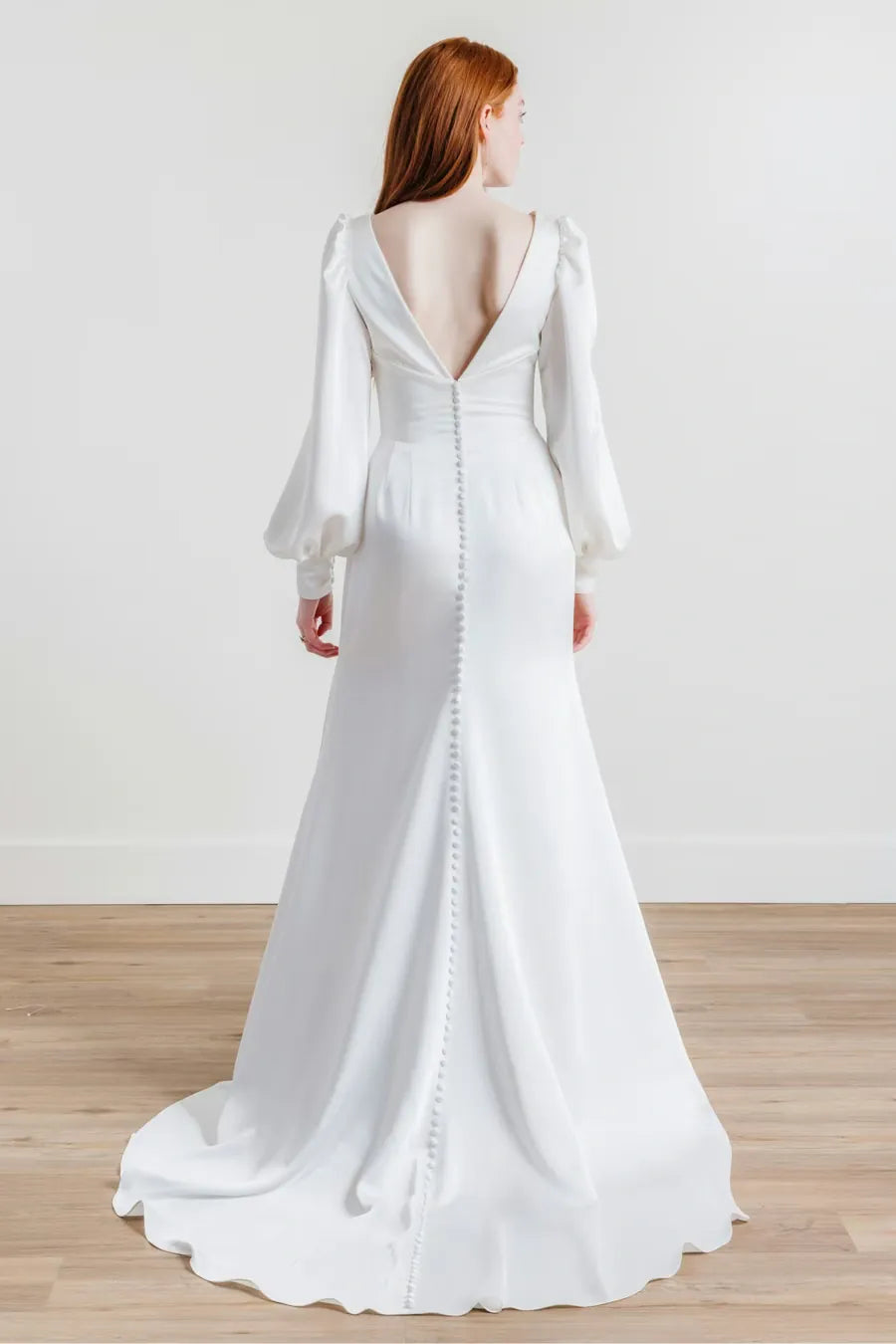 A woman wearing a white wedding dress is shown from the back in a studio setting with a light wall and wood floor. The dress has a deep V back, long balloon sleeves, and a train with decorative bead detailing running down the center.