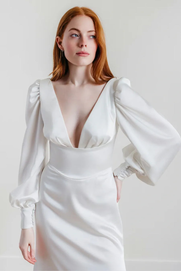 A woman with red hair wearing a white satin dress with dramatic puffed long sleeves, a deep V-neckline, and a fitted waist, photographed against a plain light background.
