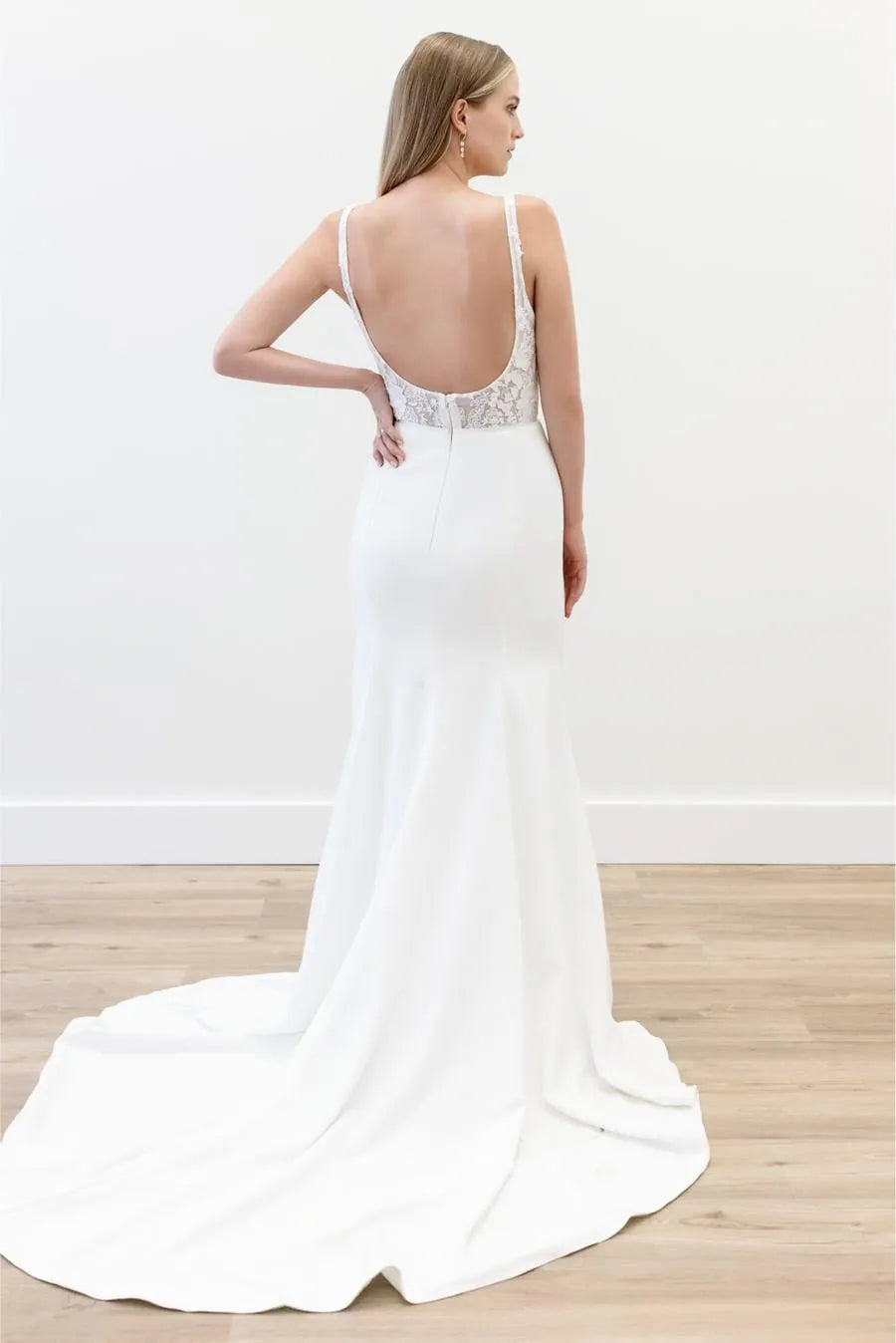 A woman modeling a white bridal gown from the back, showing a deep open back, lace-trimmed waist, and a long train on a light wood floor against a white wall.