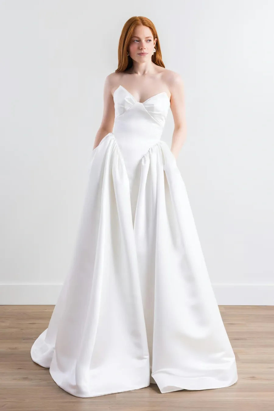 A woman modeling a strapless white wedding gown with a sweetheart neckline, gathered bodice, and full satin-like skirt on a light studio background with wood flooring.