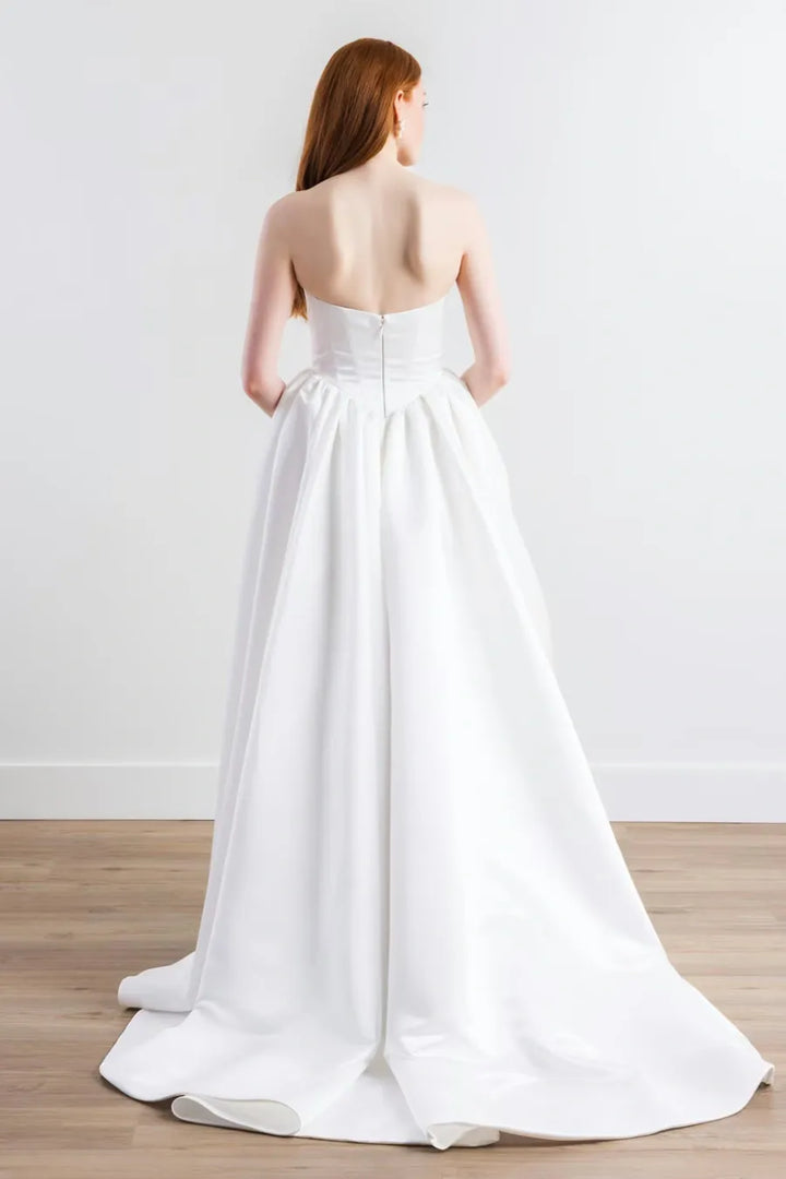 A woman shown from the back wearing a strapless white wedding gown with a structured bodice and full skirt, standing on a light wood floor against a plain white wall.